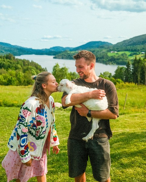 Dame og mann foran et åkerlandskap med et stort vann i bakgrunnen, mannen holder et lam